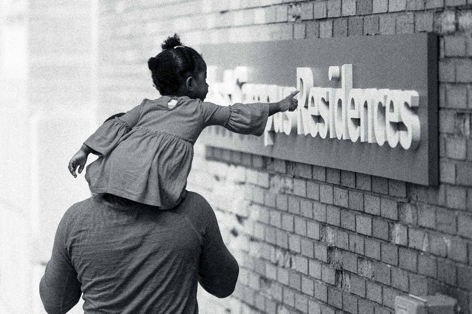 A Black little girl on an adult's shoulder's leans over to point at a Gfsntracker residence building sign